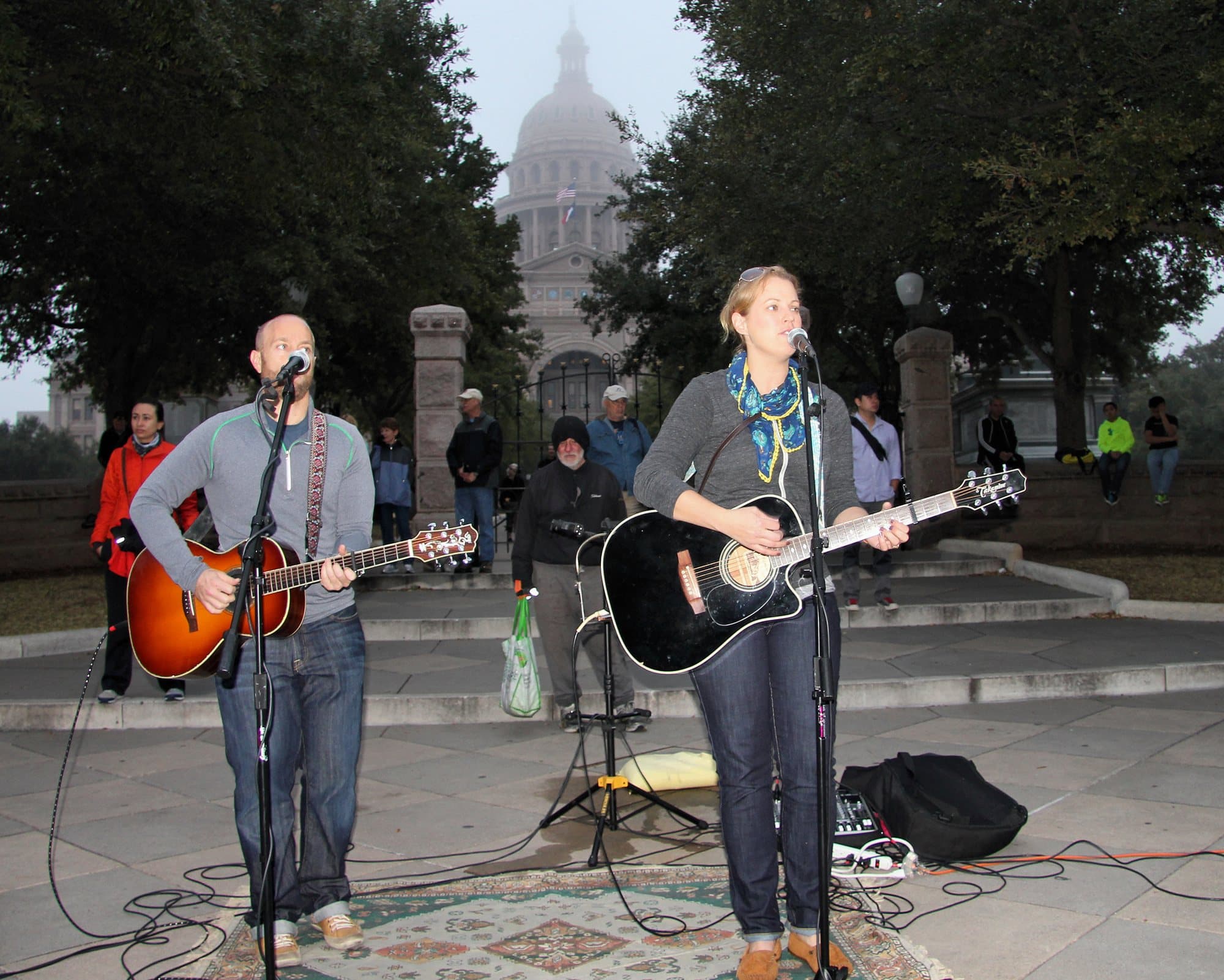 Two musicians play acoustic guitars on a stone plaza, with the Texas Capitol dome in the distance.