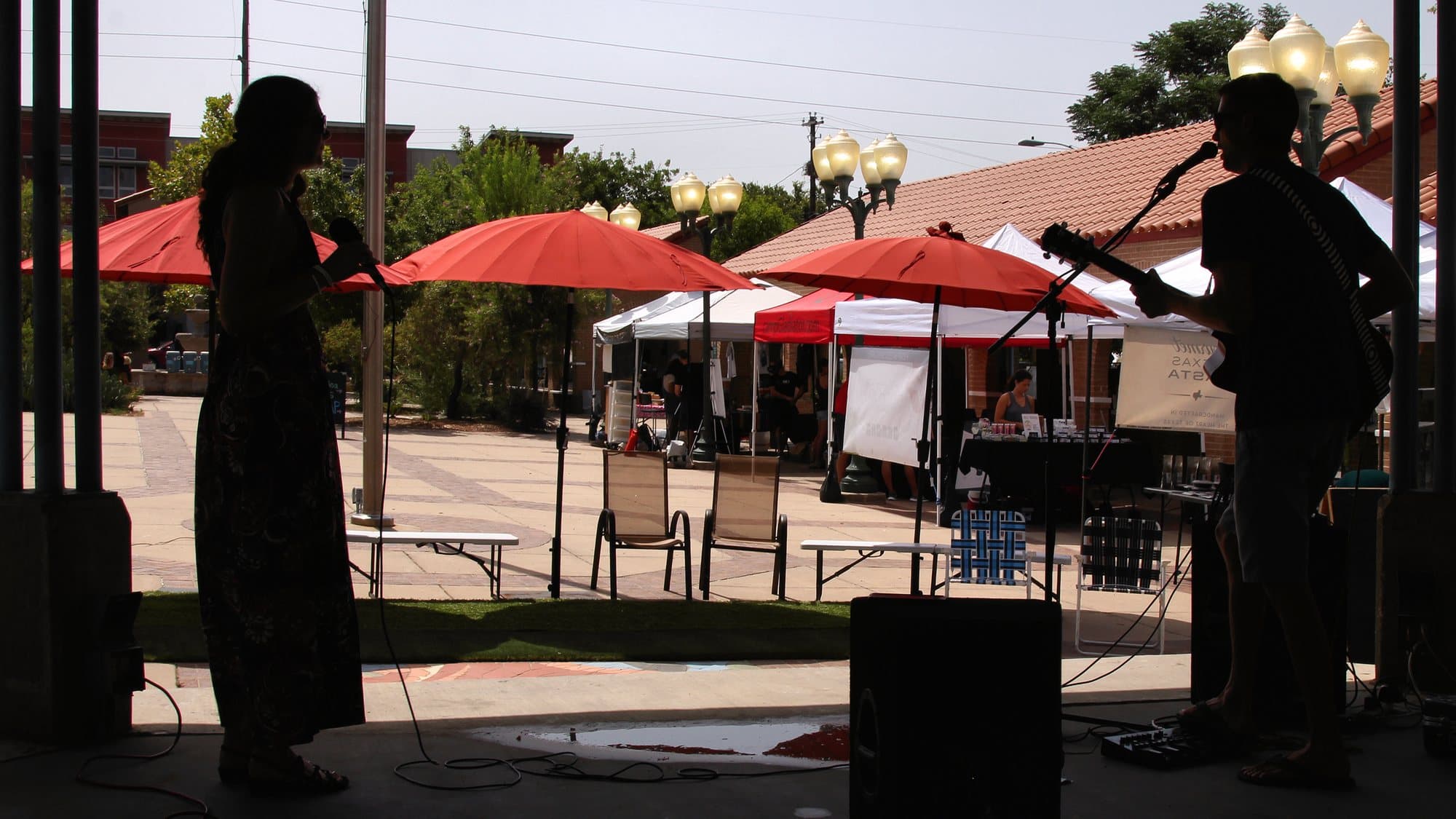 Musicians on a shaded outdoor stage, with a sunlit plaza, red umbrellas, and market tents behind them.
